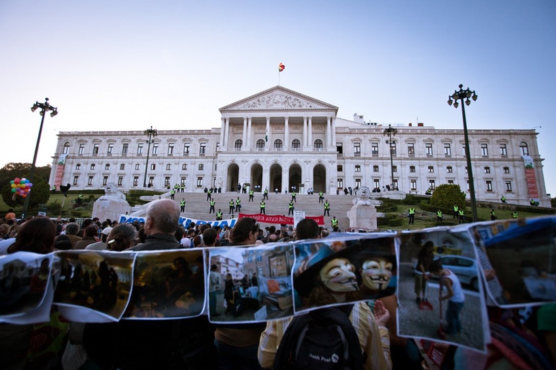 Vidvinkelvy av den portugisiska parlamentsbyggnaden, Assembleia da República, med dess neoklassiska vita fasad och kolonner. I förgrunden syns en samling demonstranter med banderoller, medan säkerhetspersonal i reflexvästar bildar en linje på trappan. Den portugisiska flaggan vajar från byggnadens topp.