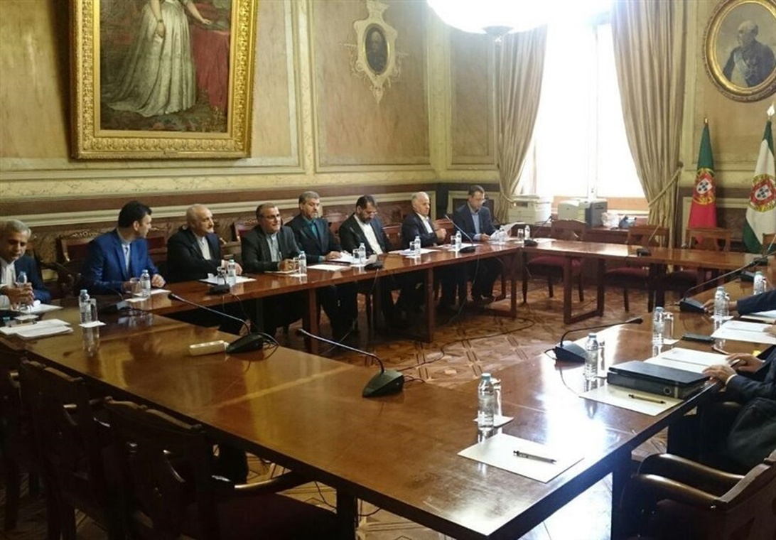 Interiörvy av ett mötesrum i det portugisiska parlamentet där tjänstemän sitter vid ett långt träbord för vad som verkar vara ett kommittémöte eller diplomatisk diskussion. Rummet har elegant inredning med historiska målningar, utsmyckade guldramar och portugisiska flaggor synliga i bakgrunden.