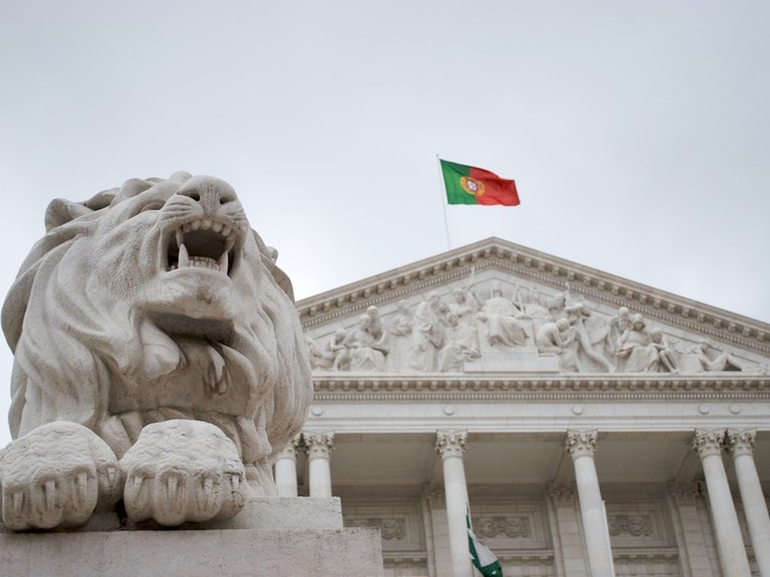 En slående komposition som visar fasaden på den portugisiska parlamentsbyggnaden (São Bento-palatset) med dess klassiska arkitektur, kolonner och triangulära pediment. I förgrunden finns en av de ikoniska stenlejonstatyerna som vaktar parlamentet, medan den portugisiska flaggan vajar framträdande från toppen av byggnaden mot en grå himmel.